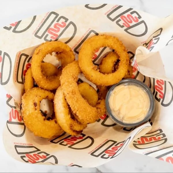 Golden-brown onion rings and a dipping sauce cup in a Wagyu Burger Shack branded paper-lined basket.
